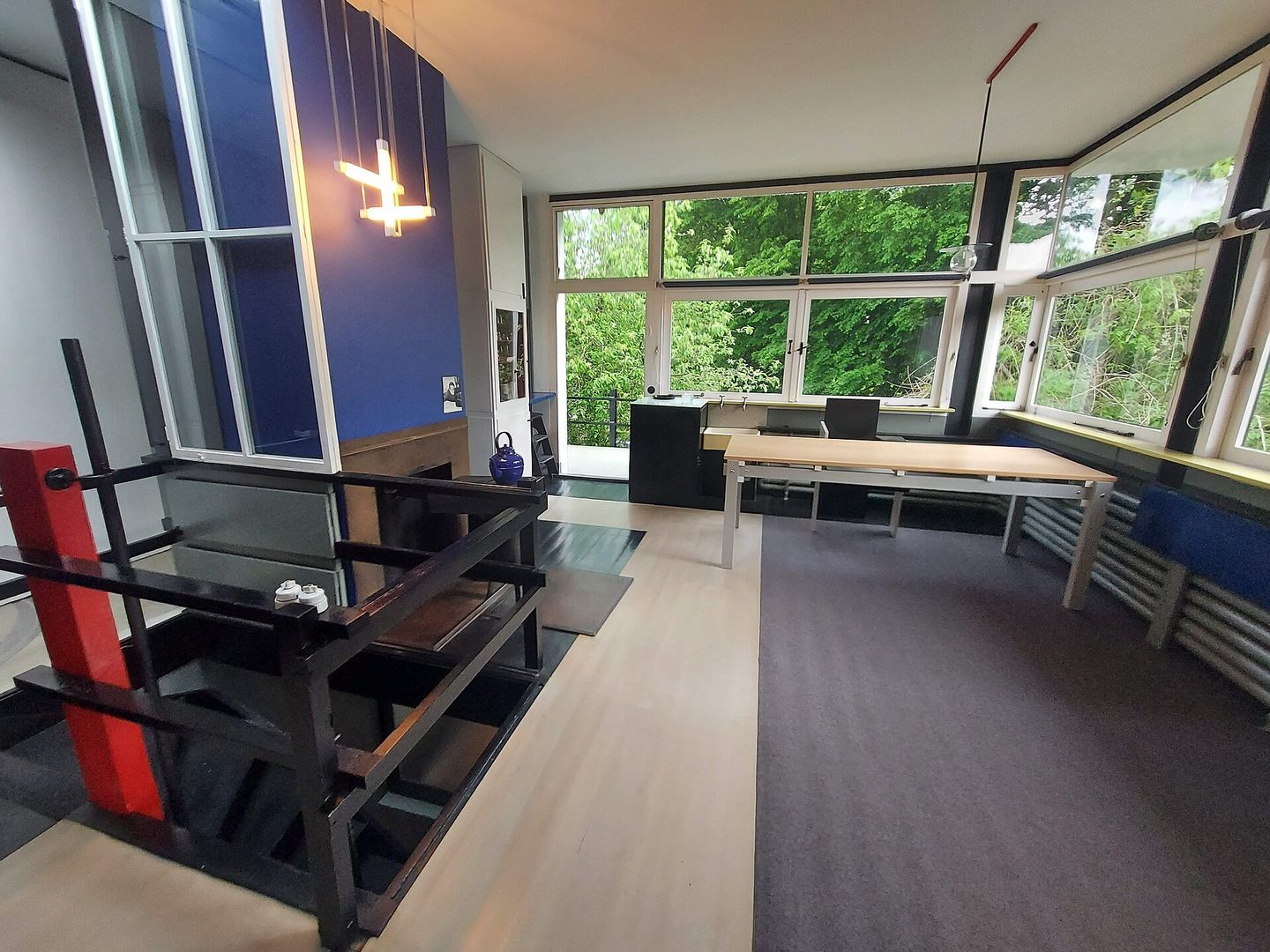 Lichte kamer met grote hoekramen, een houten tafel, zwarte stoelen en een rood-zwart traphek