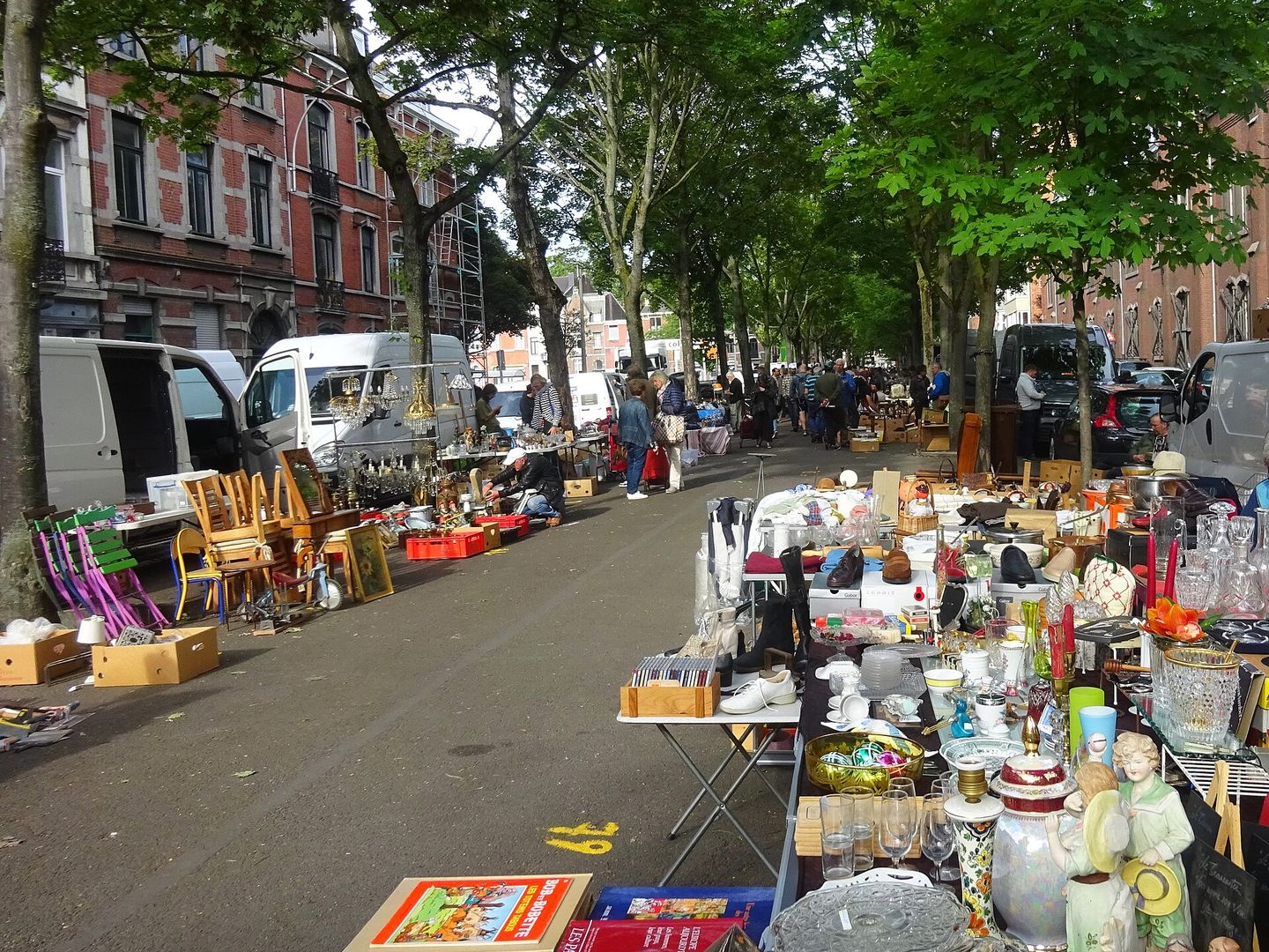 Tafels vol servies, beelden, boeken en stoelen langs een straat met bomen