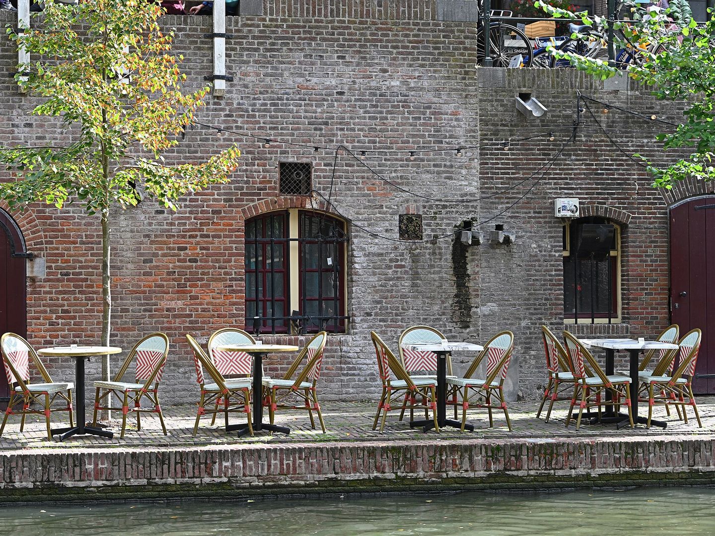 Terras met bistrostoelen op een werf langs de Oudegracht, voor een bakstenen gevel met boogdeuren