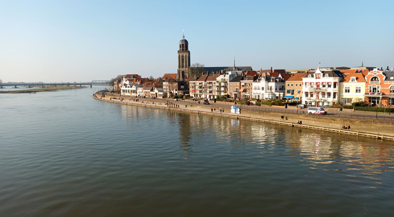 De IJsselkade met een rij huizen en de toren van de Lebuinuskerk, gezien over het water van de IJssel