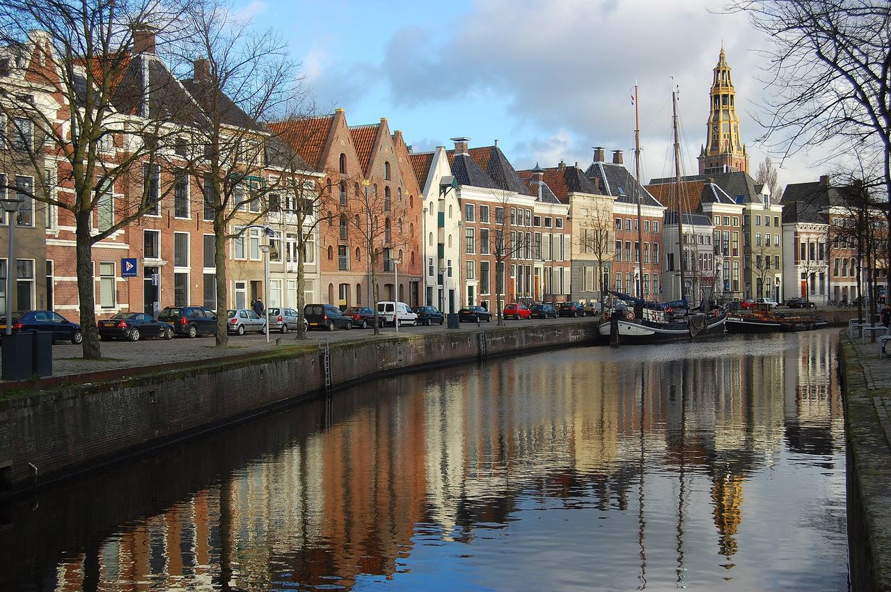 Pakhuizen en grachtenpanden aan de Hoge der A, gespiegeld in het water, met de toren van de Der Aa-kerk op de achtergrond