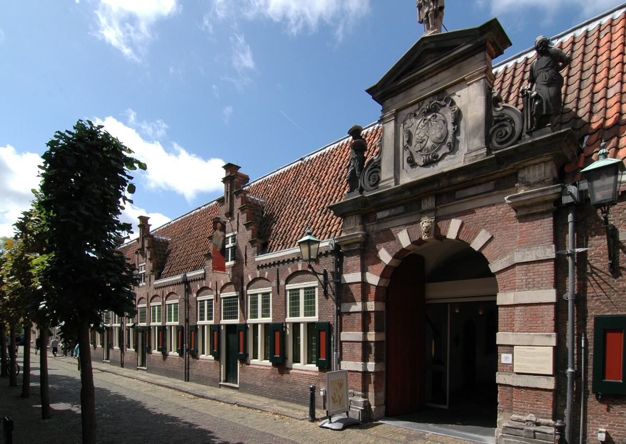 Toegangspoort van het voormalige Oudemannenhuis met beelden op de gevel en een lange rij lage huisjes