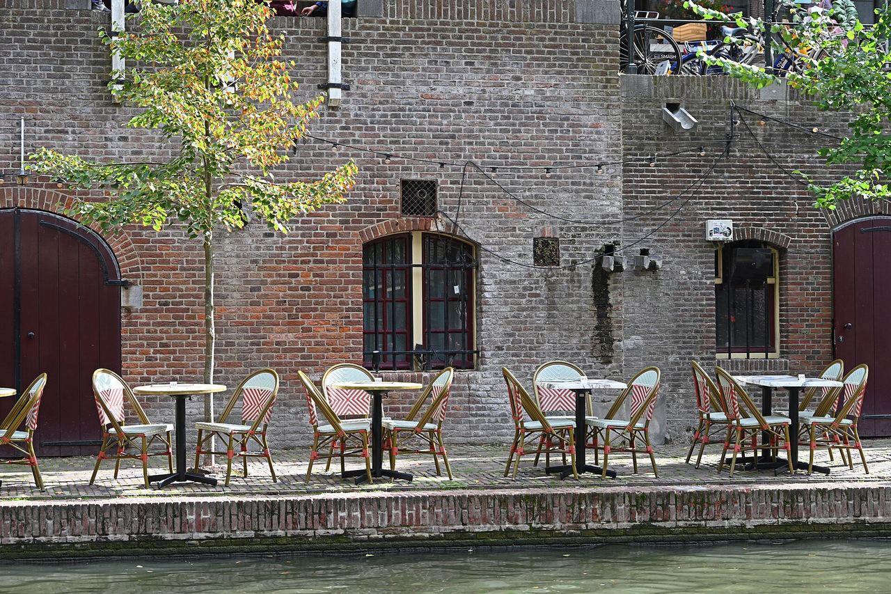 Terras met bistrostoelen op een werf langs de Oudegracht, voor een bakstenen gevel met boogdeuren