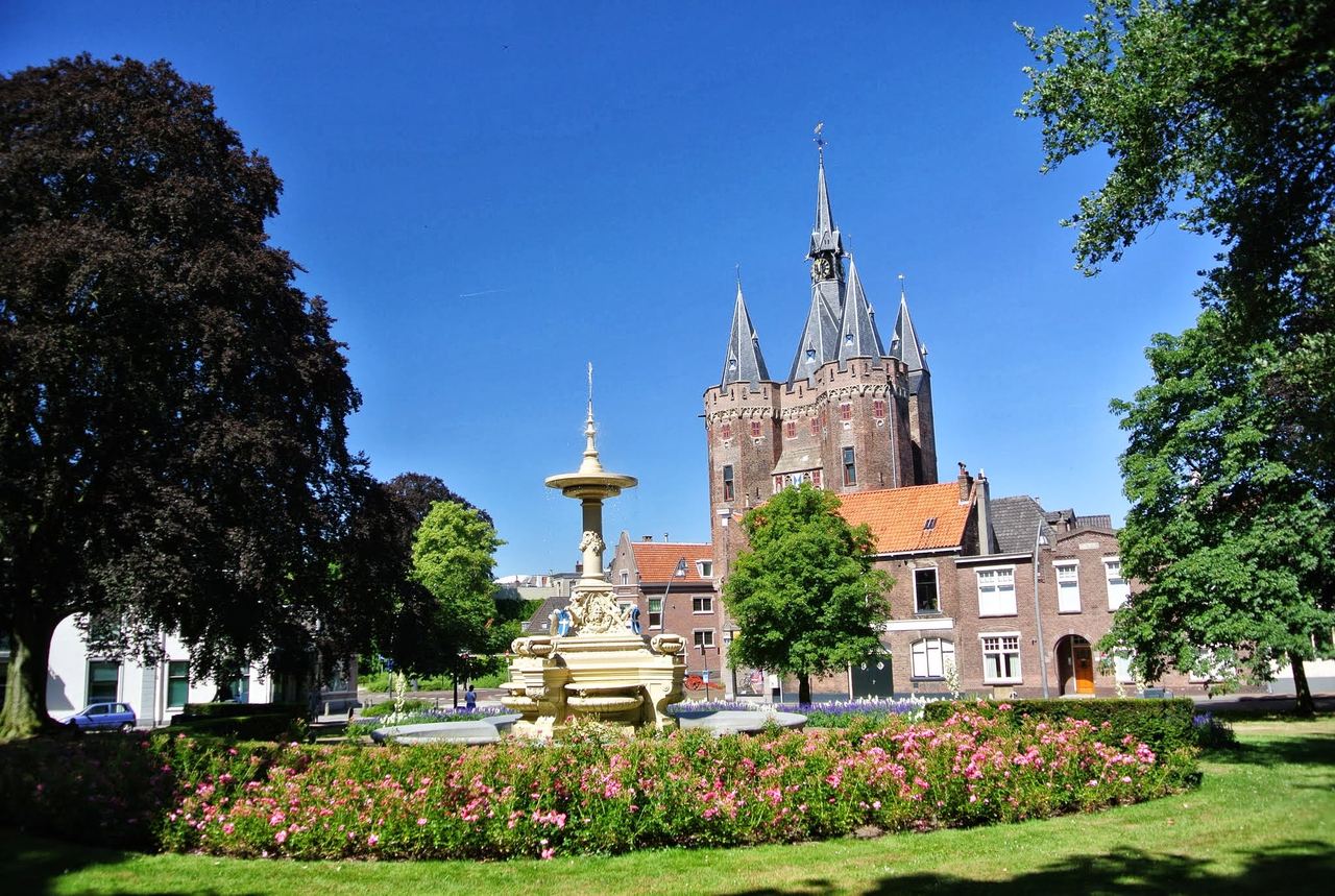 De Sassenpoort met spitse torentjes achter een plantsoen met bloemen, een fontein en hoge bomen