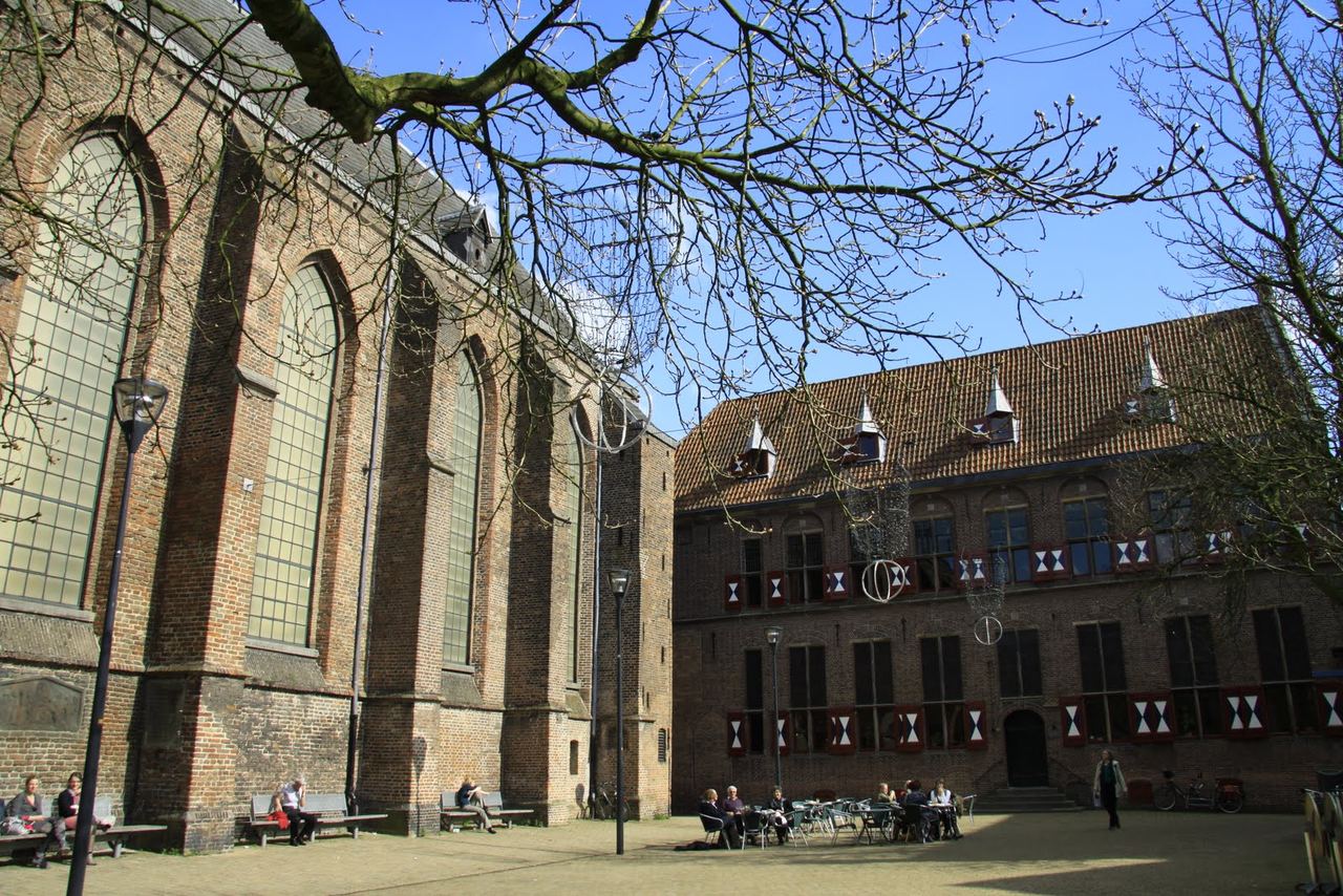 Hoge bakstenen kerkgevel met spitsboogramen naast de refter met rood-witte luiken, aan een plein met terrassen