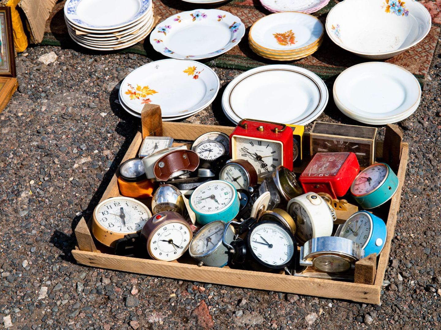 Houten krat vol oude wekkers naast stapels borden op een vlooienmarkt