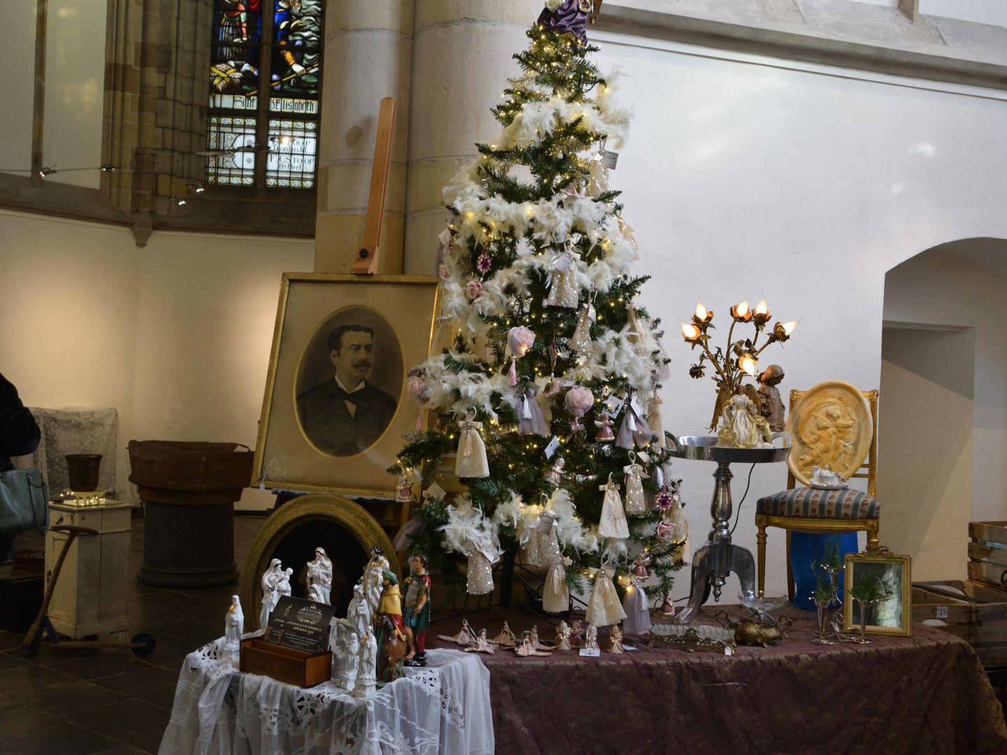 Kraam met kerstbrocante in de kerk: een witte kerstboom, engeltjes, kandelaars en een ingelijst portret