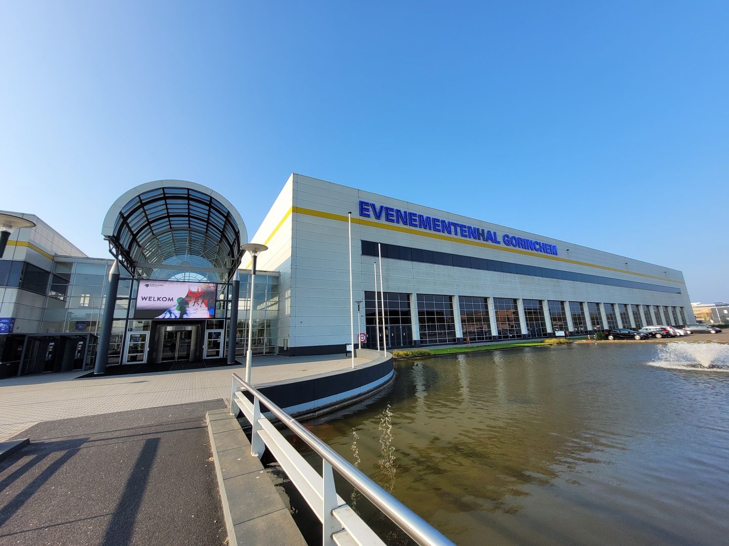 Evenementenhal Gorinchem met de glazen entreekoepel aan een vijver onder een blauwe lucht