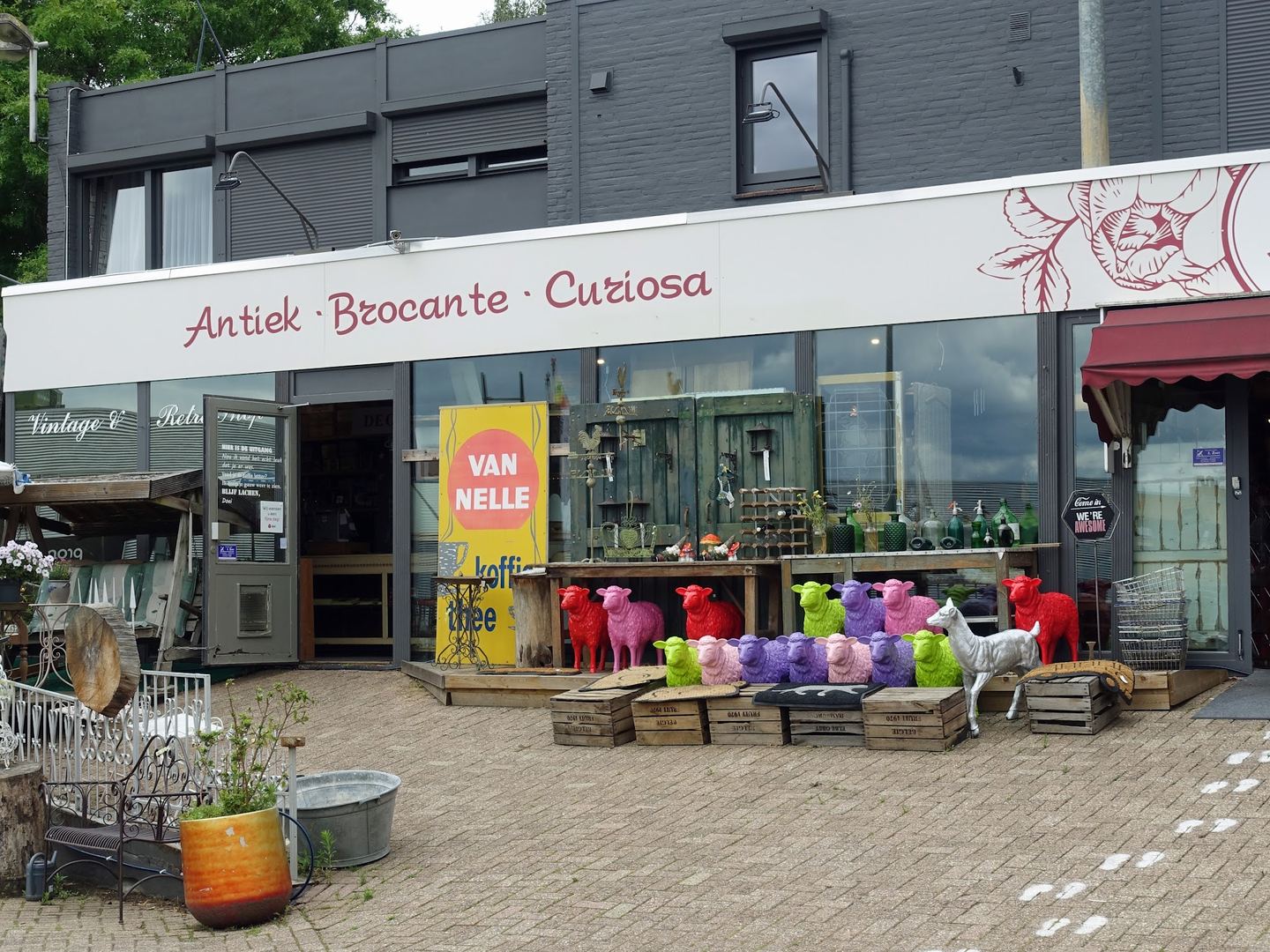 Gevel van Adelheid Antiek, Brocante en Curiosa met tuinmeubels en gekleurde schapenbeelden voor de deur