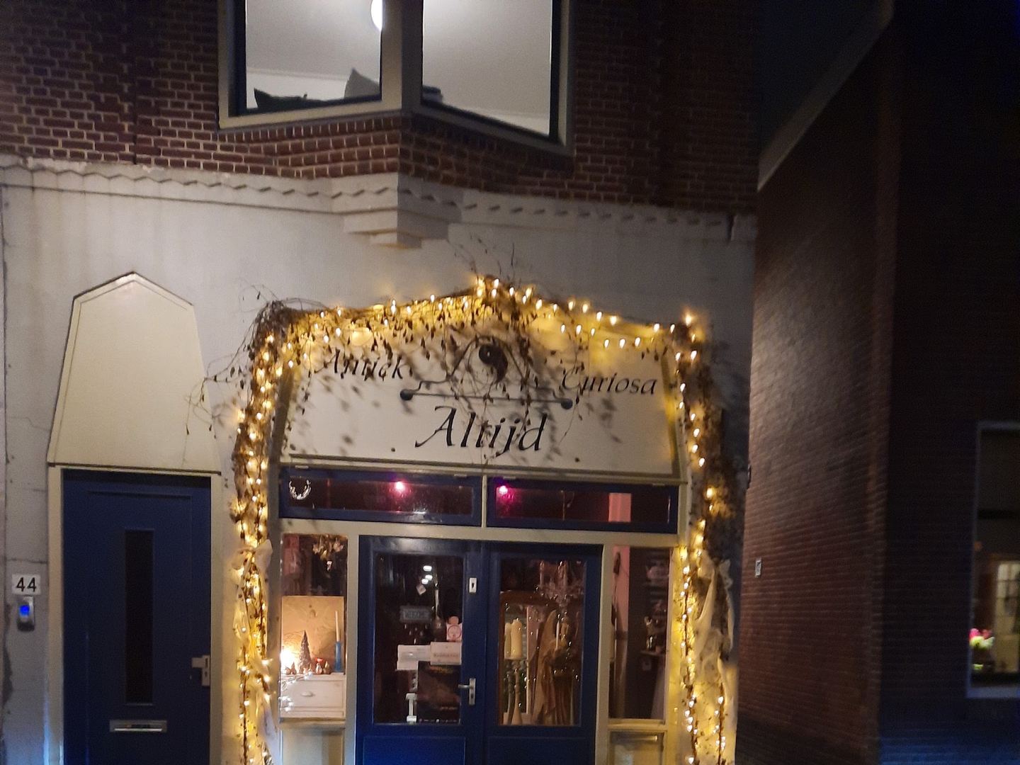 Winkelpui van Antiek Curiosa Altijd bij avond, met naambord en blauwe deuren omlijst door lichtjes