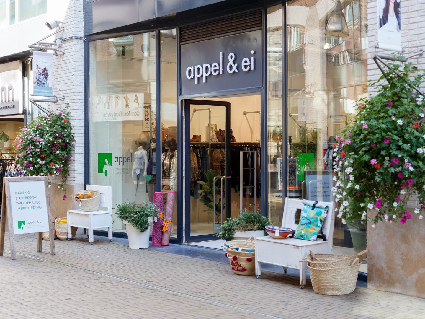 Glazen winkelpui van Appel & Ei met naambord boven de deur, hangende bloemen en bankjes met manden op de stoep