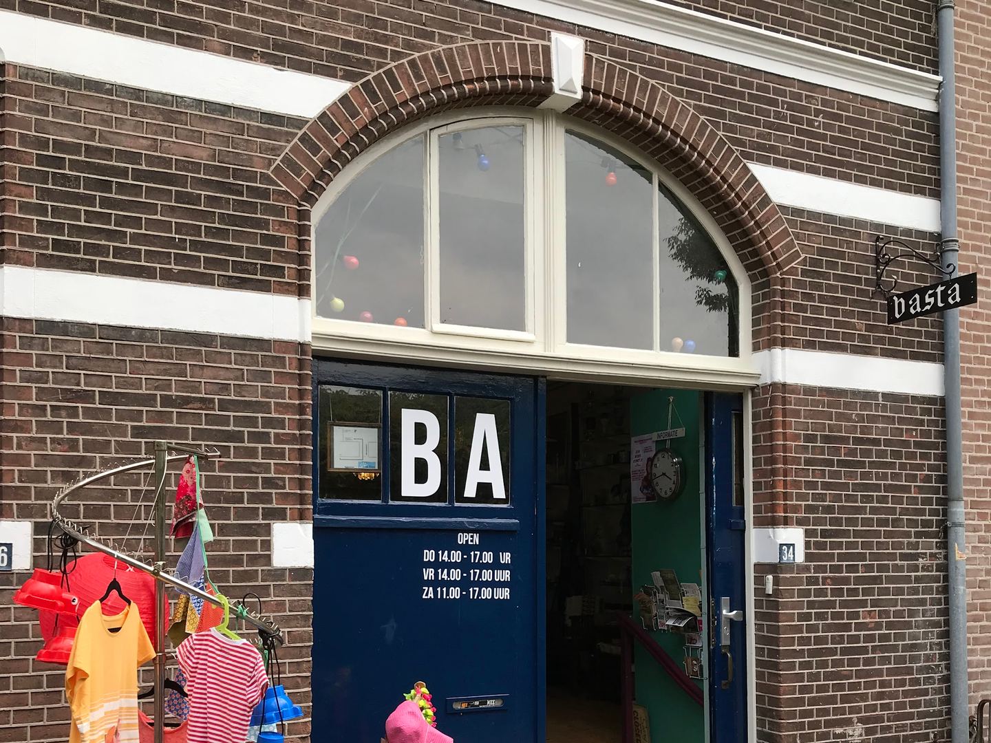Bakstenen gevel van Basta met de blauwe toegangsdeur en kleding aan een rek voor de deur