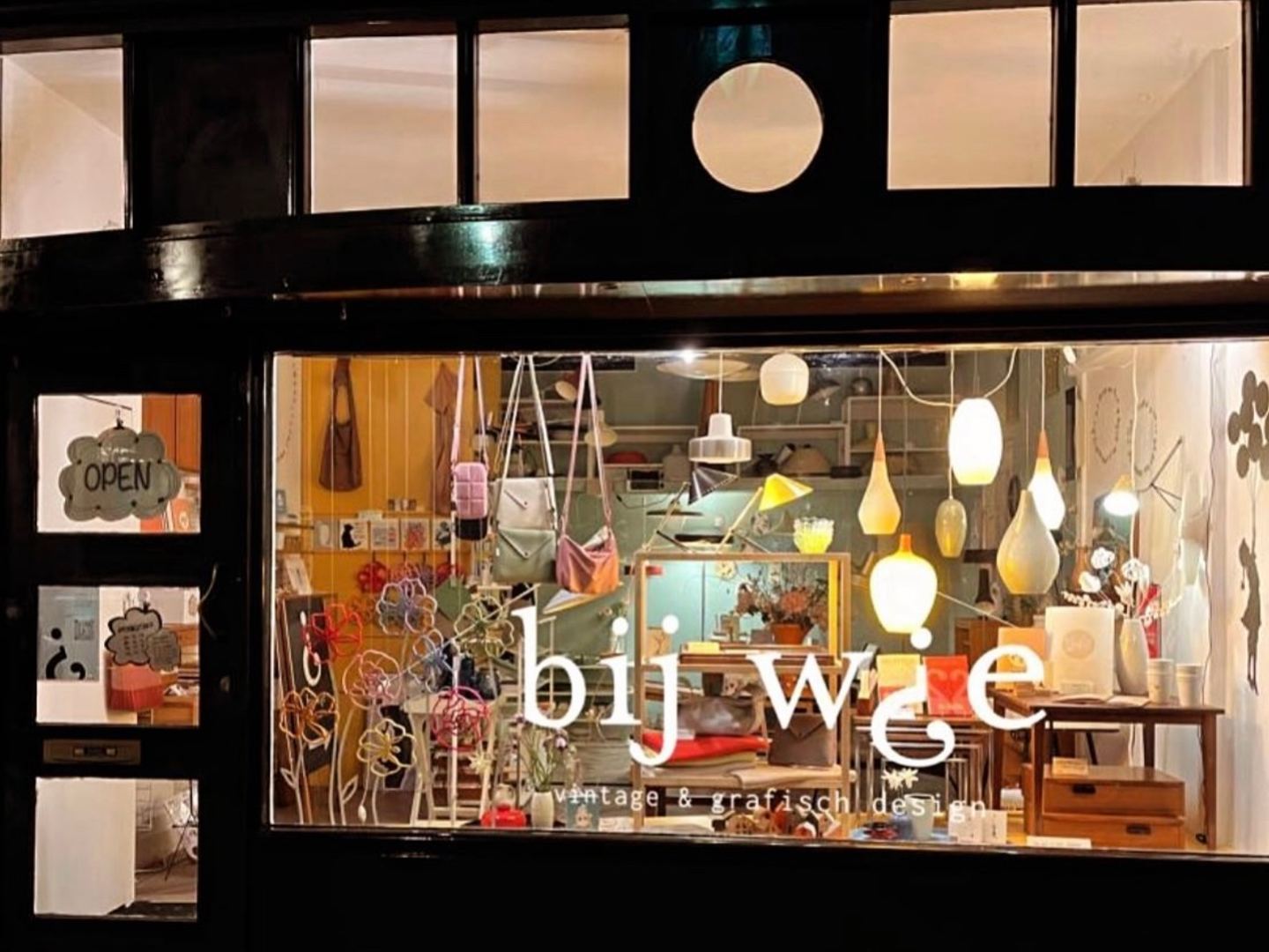 Verlichte etalage van Bij Wie met vintage hanglampen, tassen en meubels, bij avond