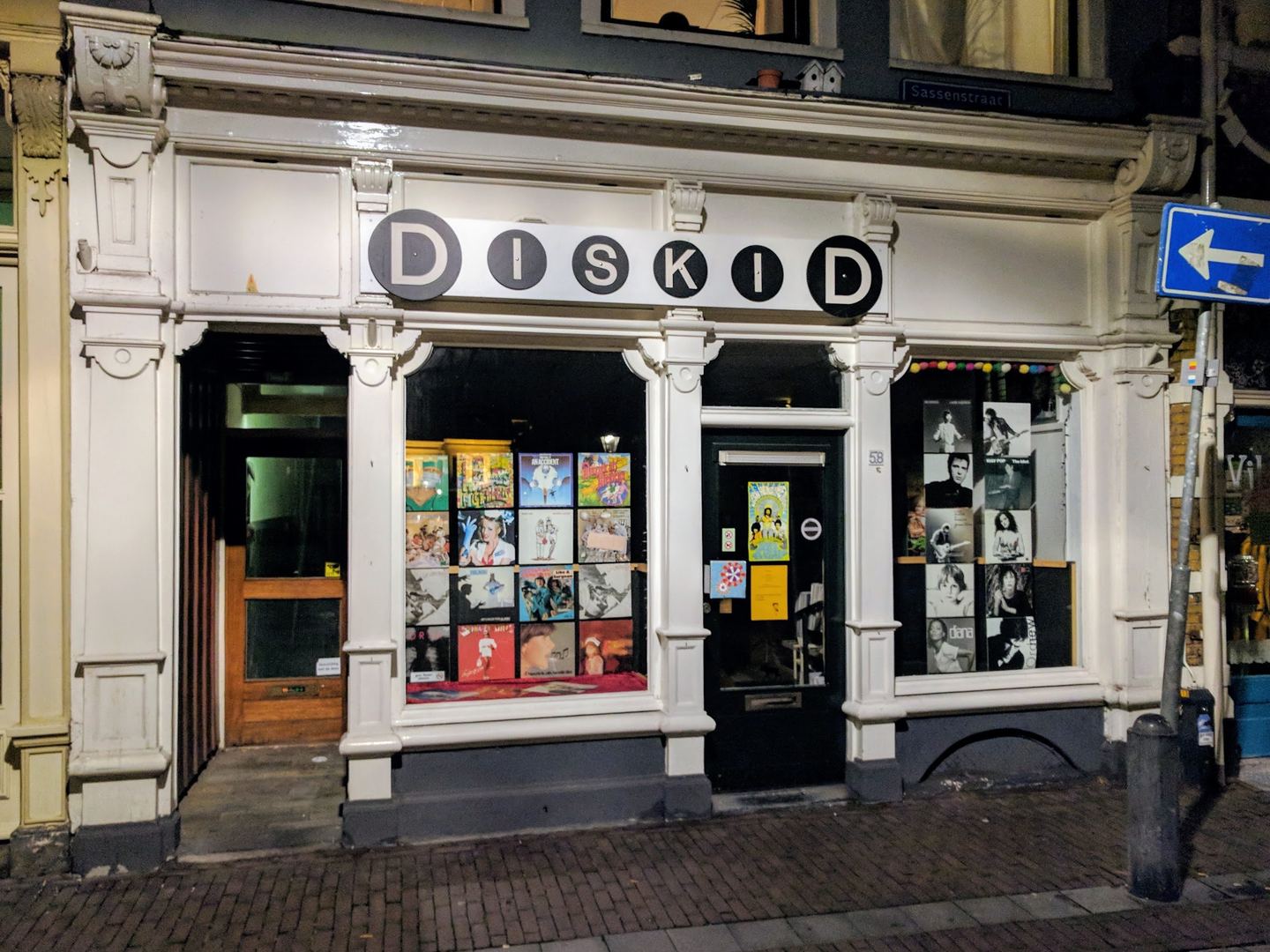 Witte winkelpui van Diskid met zwart-wit naambord en verlichte etalages vol platenhoezen in de avond