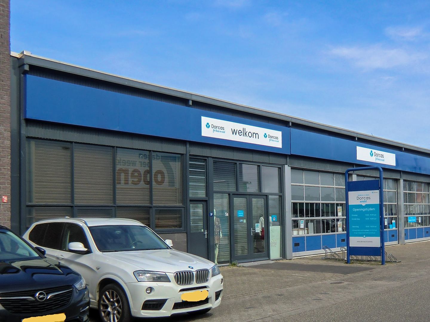 Blauwe gevel van de Dorcas-kringloopwinkel met welkomstbord en vlaggen bij de ingang