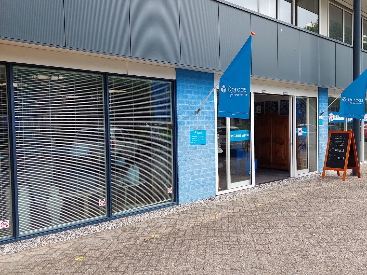Ingang van Dorcas Winkel Veenendaal met blauwe Dorcas-vlaggen en een stoepbord