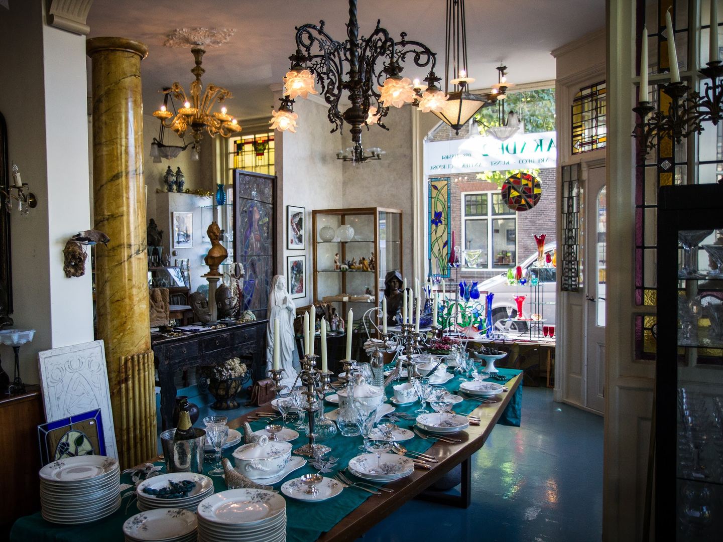 Kroonluchters, servies en glaswerk op tafels in de winkel van Emmakade Antiek