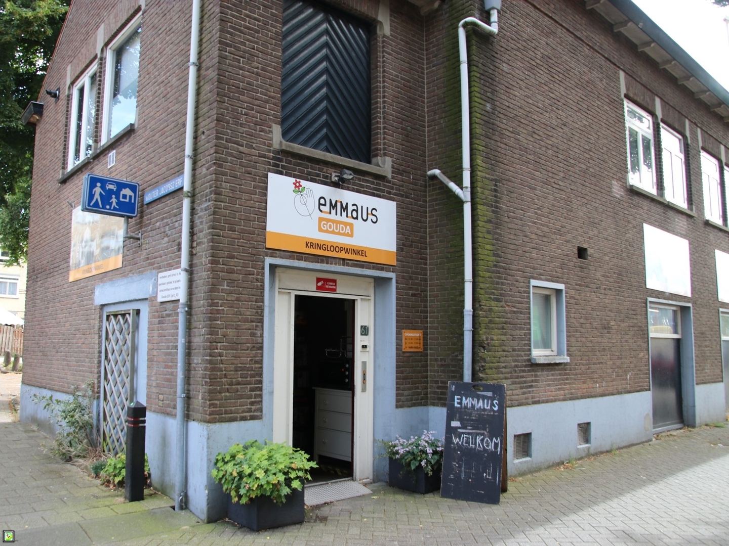 Bakstenen gebouw van Emmaus Gouda met oranje kringloopbord boven de ingang en een welkomstbord