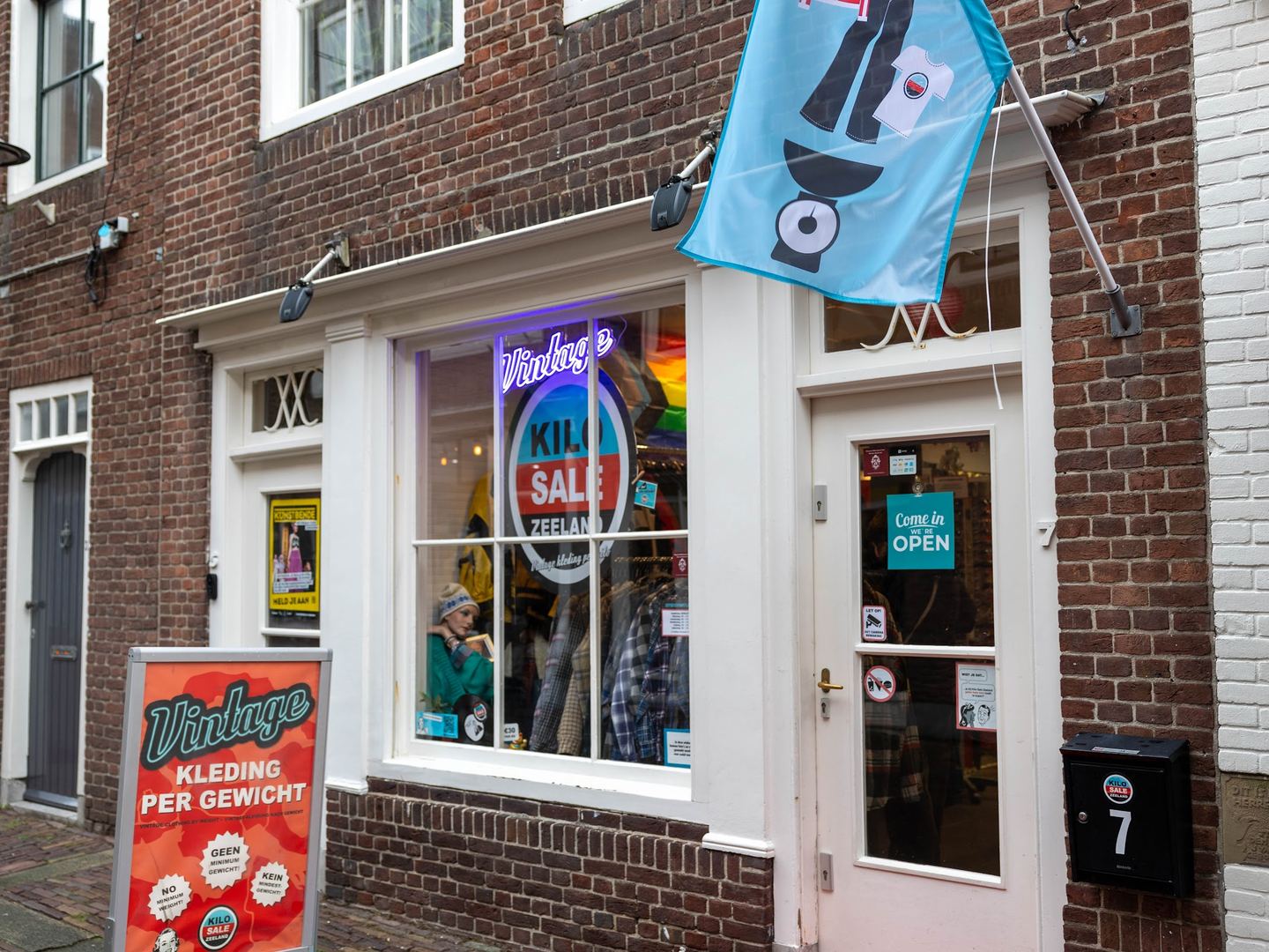 Gevel van Kilo Sale Zeeland met neonlogo in de etalage en een stoepbord 'Vintage kleding per gewicht'