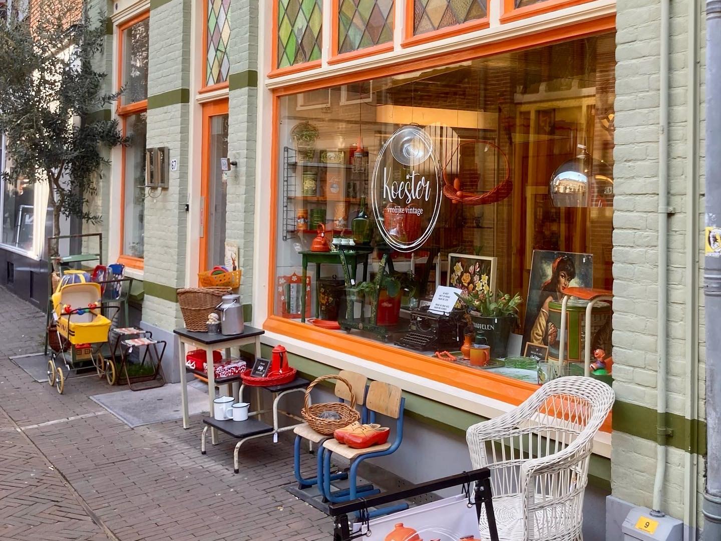 Gevel van Koester met oranje kozijnen, glas-in-loodramen en een etalage met vintage spullen op de stoep