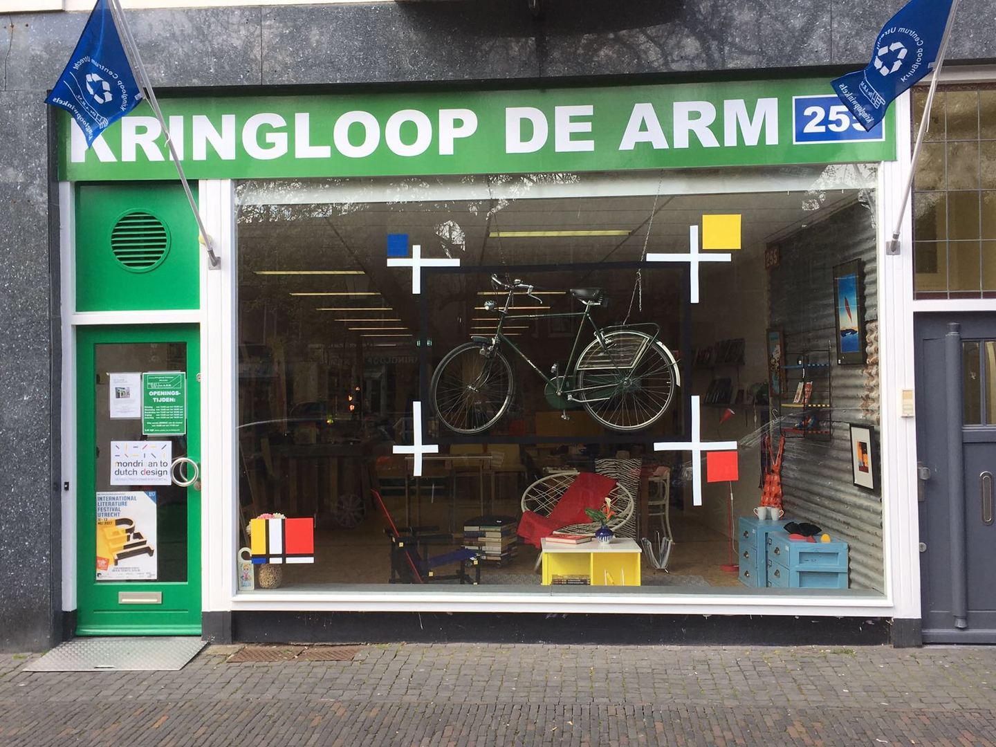 Groene winkelpui van Kringloop De ARM met naambord en een etalage met een fiets en meubels