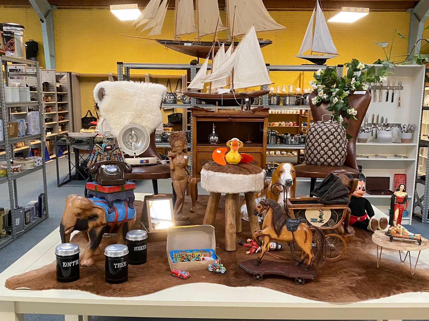 Interieur van Kringloopwinkel Alkmaar met een tafel vol curiosa, modelzeilschepen en schappen met huisraad