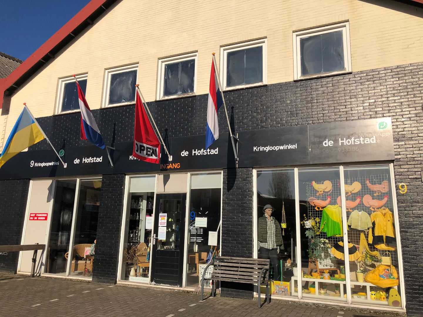 Gevel van Kringloopwinkel De Hofstad met vlaggen en volle etalages