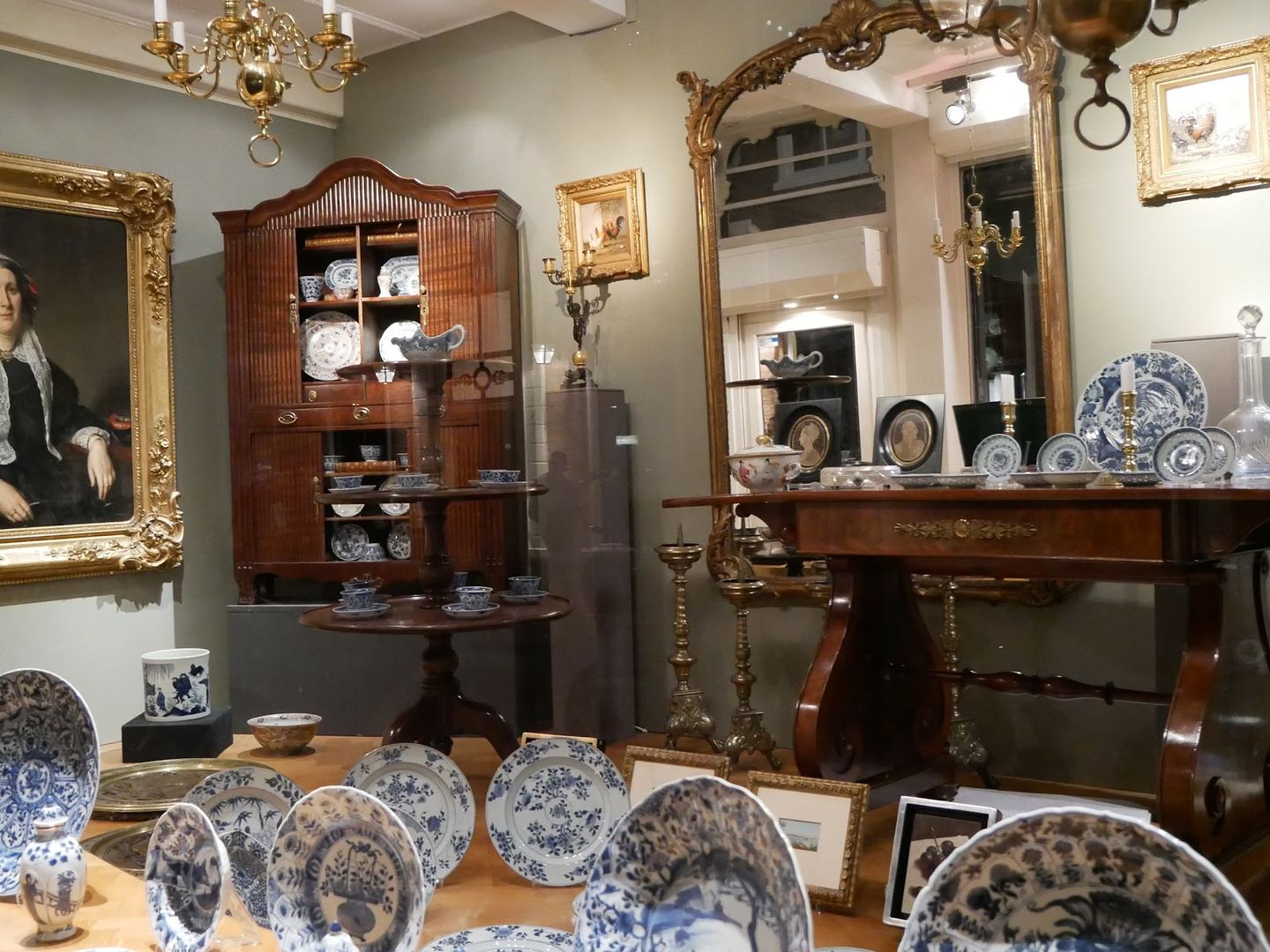 Interieur van Limburg Antiquairs met een geschilderd portret, een antieke kast, klokken en blauw-wit porselein op tafels