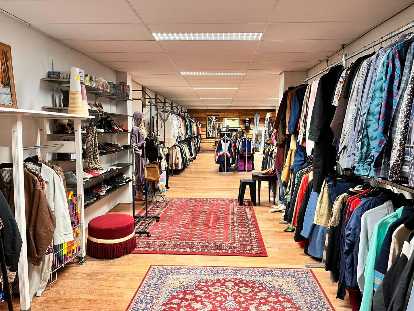Winkelvloer van MoDa met rekken vol tweedehands kleding, schoenen op planken en rode tapijten op de houten vloer