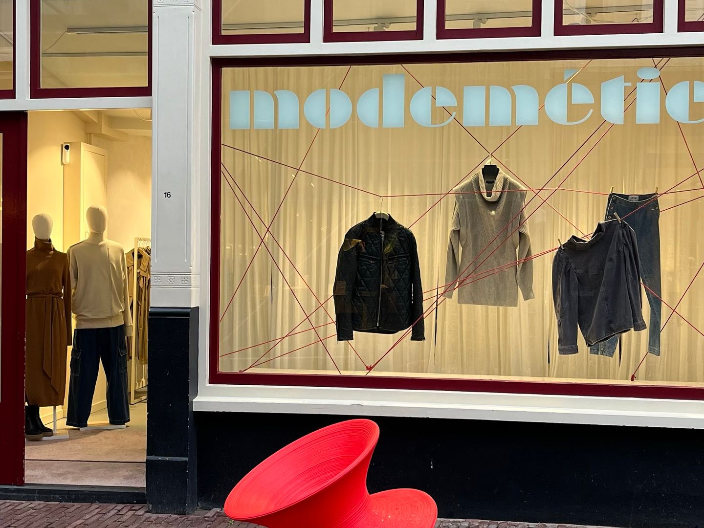 Etalage van Modémétier met de winkelnaam op het raam en kleding achter rode draden