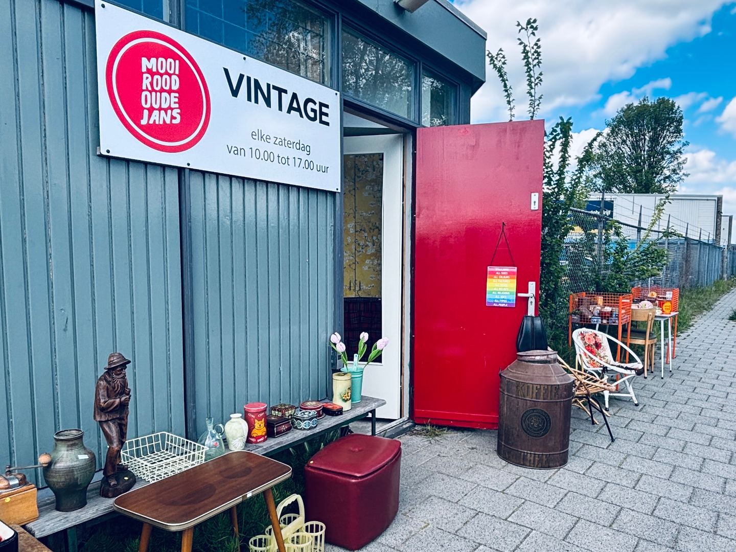 Ingang van Mooi Rood Oudejans Vintage met naambord, rode deur en vintage meubels en vazen buiten