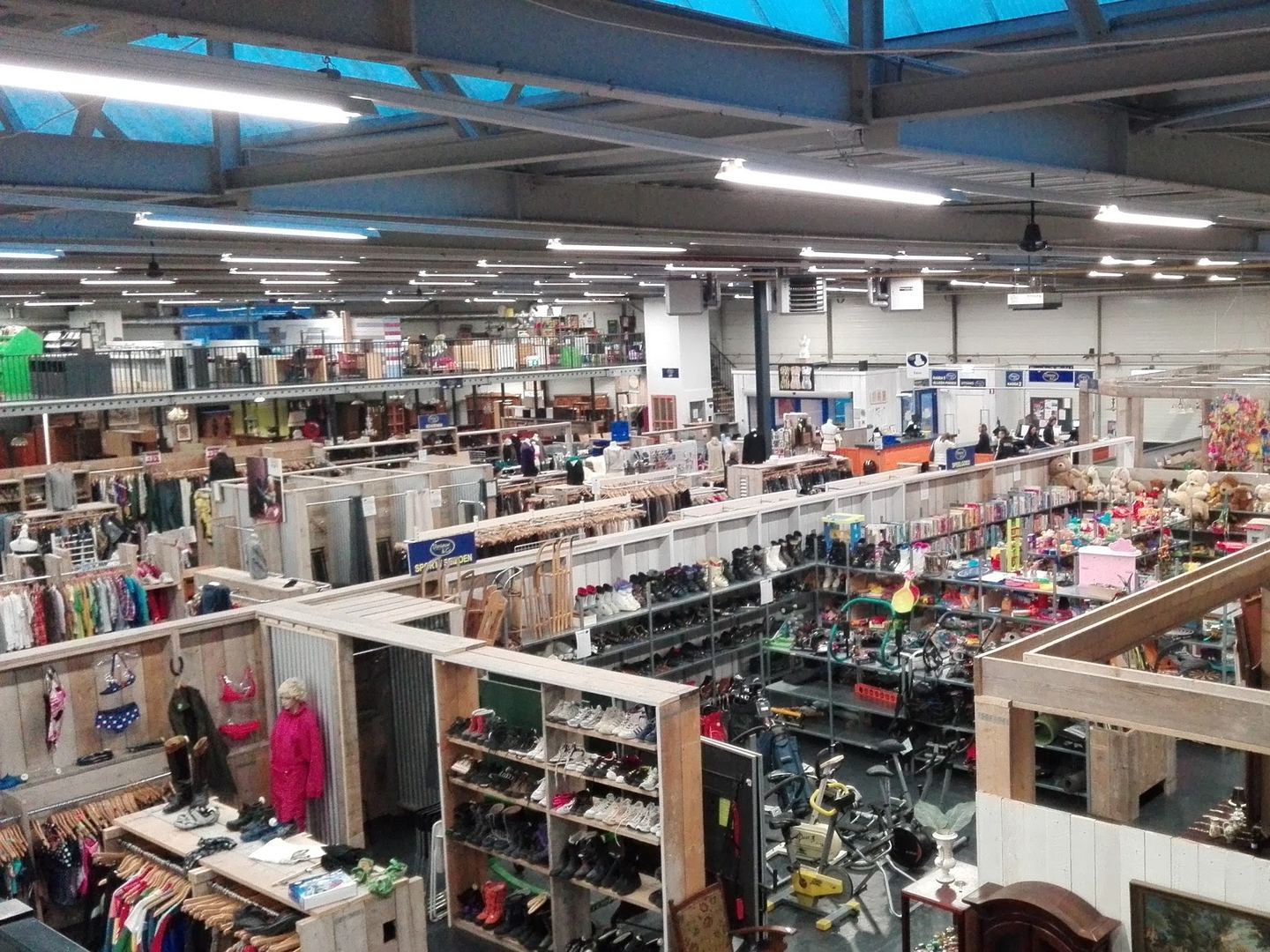 Overzicht van de grote kringloophal met afdelingen voor kleding, schoenen, speelgoed en huisraad