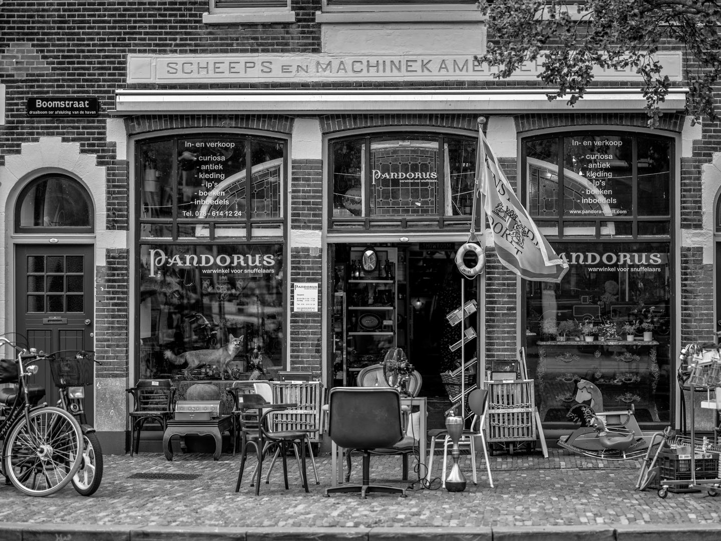 Zwart-witfoto van de bakstenen gevel van Pandorus met stoelen, rekken en curiosa op de stoep