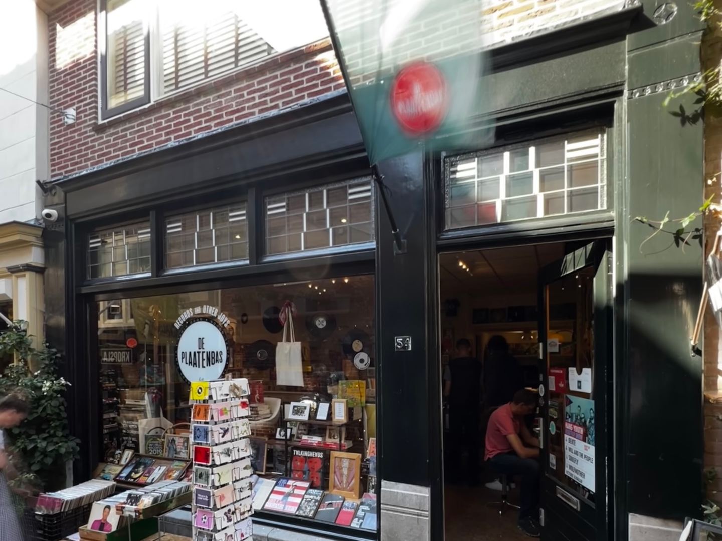 Gevel van Plaatenbas met logo in de etalage, open deur, ansichtkaartenrek en platenbakken op de stoep