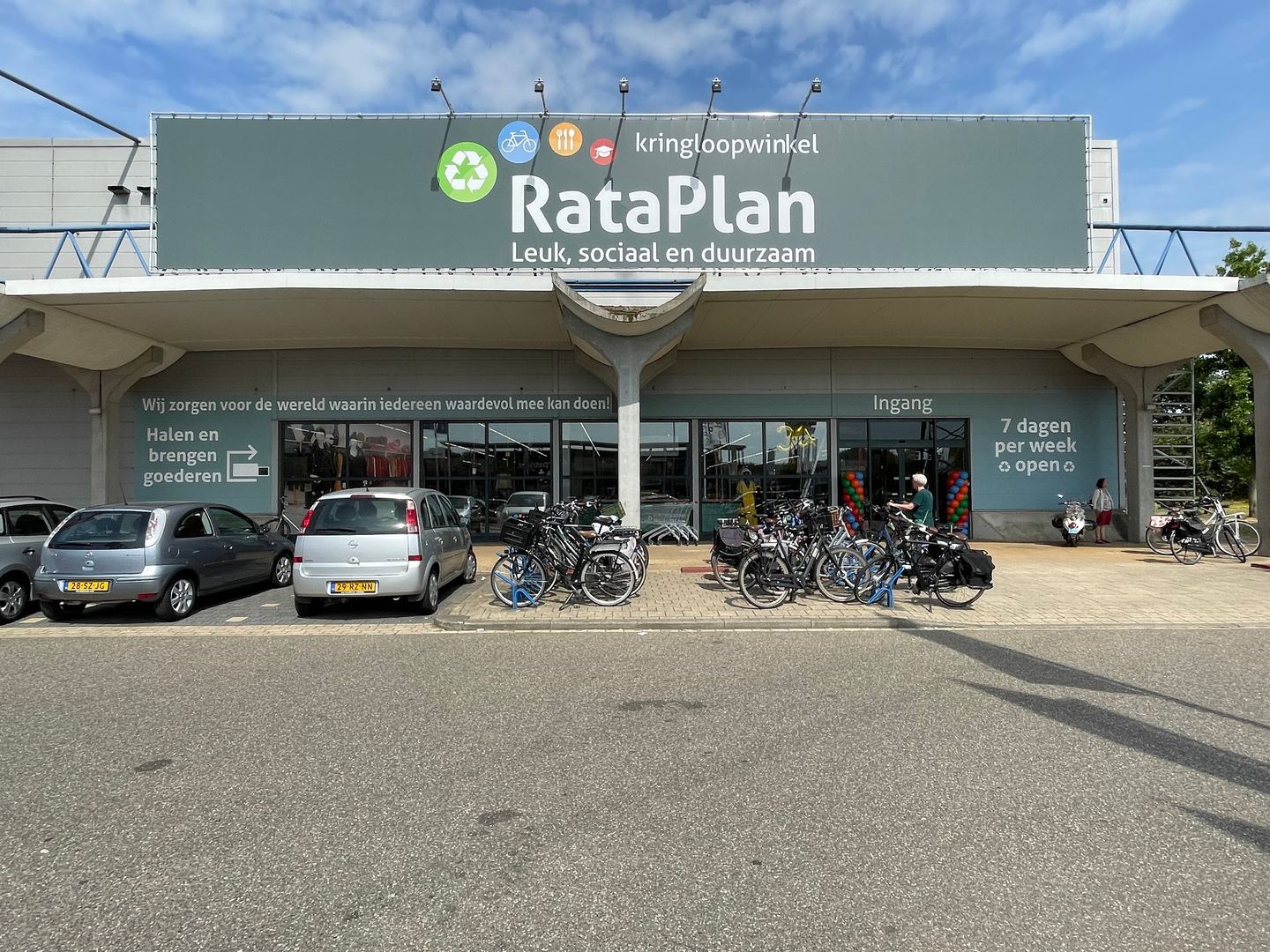Gevel van Kringloopwinkel RataPlan met groot naambord, met fietsen en geparkeerde auto's voor de ingang