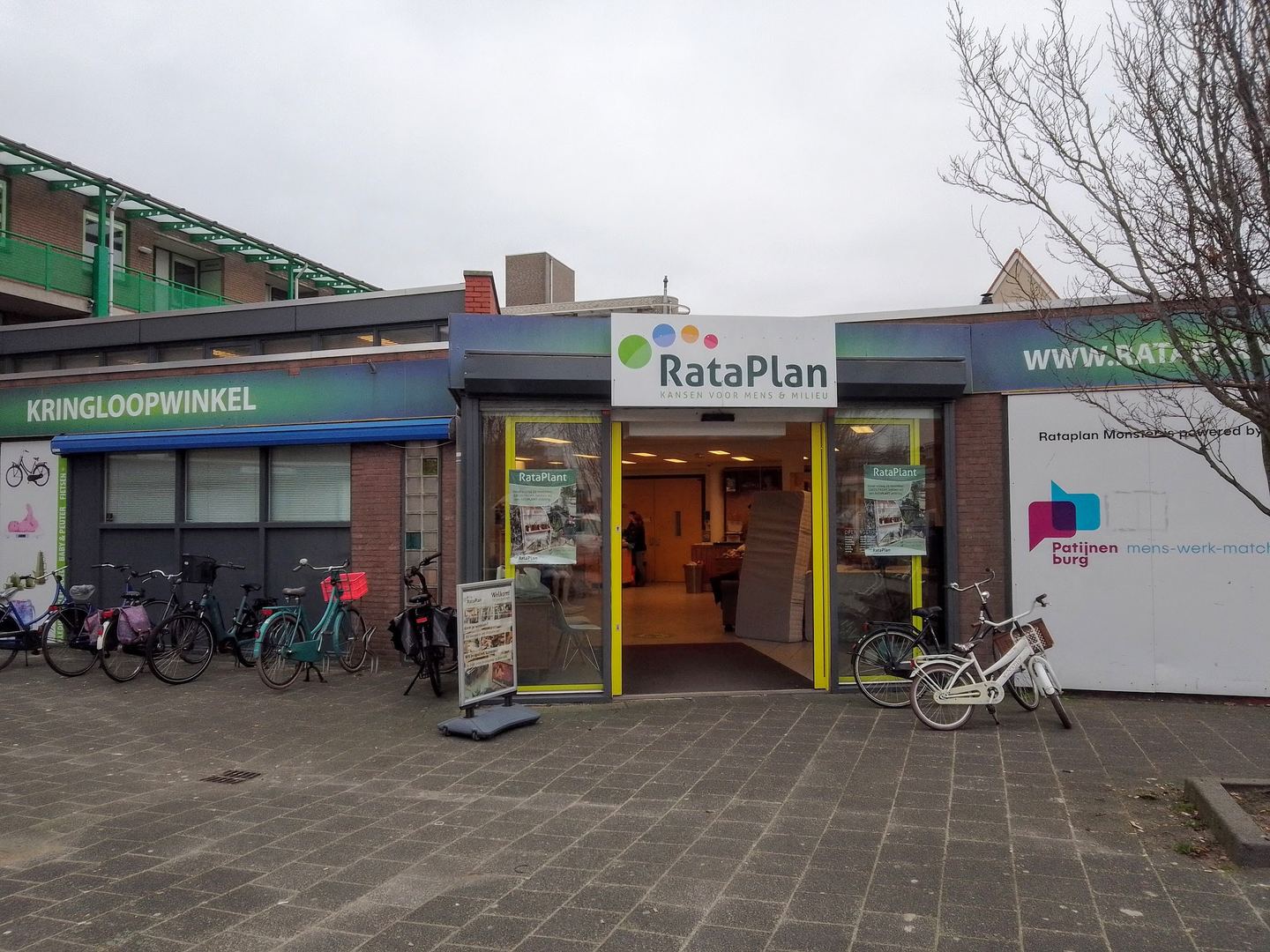 Ingang van Kringloopwinkel RataPlan Monster onder een flatgebouw, met het RataPlan-bord en fietsen voor de deur