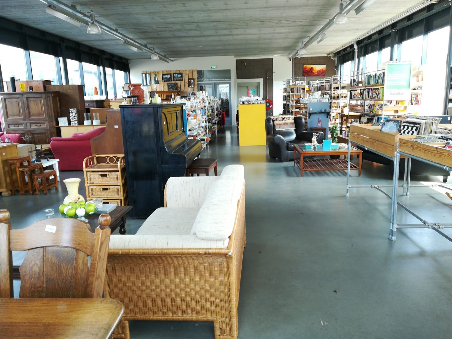Interieur van Kringloopwinkel RataPlan Naaldwijk met meubels, een piano, een rieten bank en schappen vol spullen