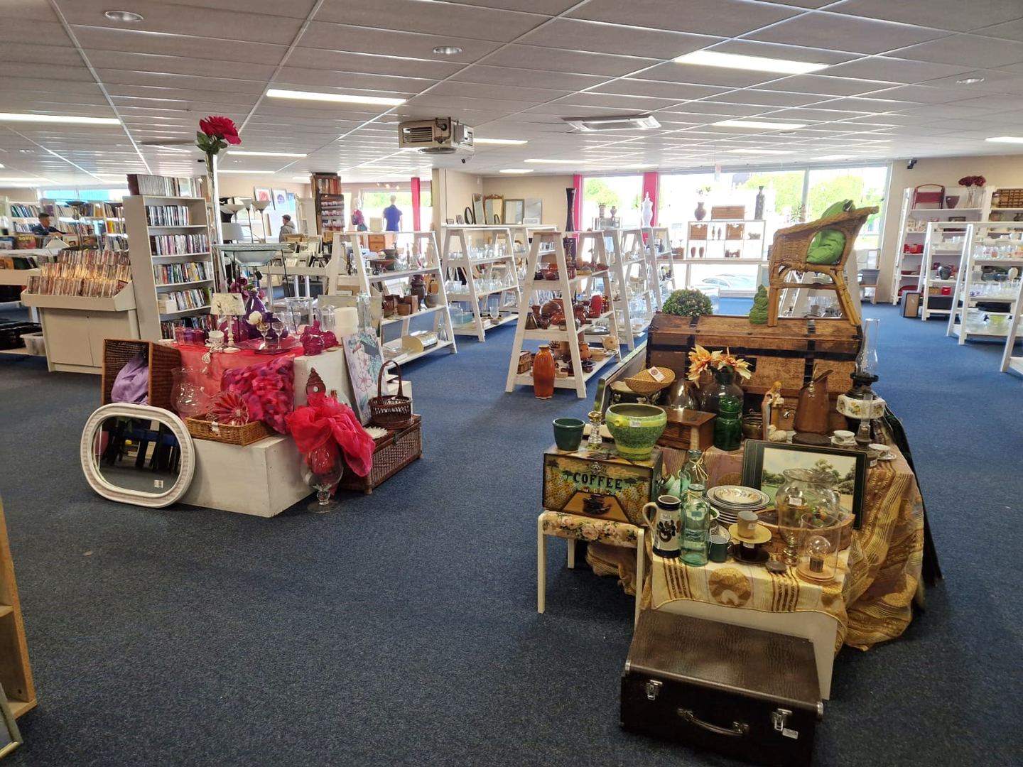 Winkelvloer van Kringloopwinkel RataPlan Velserbroek met boekenkasten, witte schappen met glaswerk en tafels vol curiosa