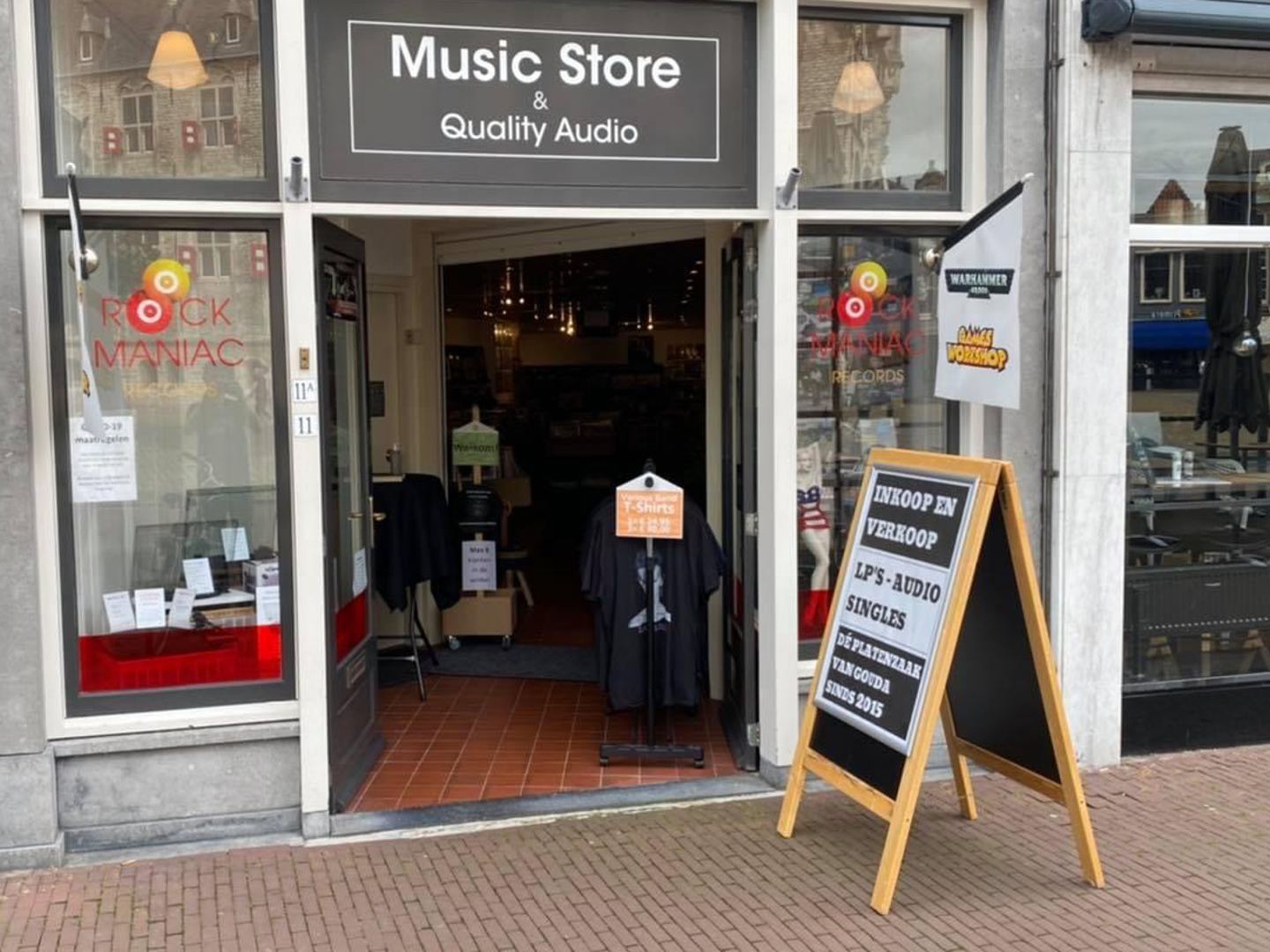 Winkelpui van Rock Maniac Records met het logo op de etalageruiten, open deur en stoepbord voor platen en audio