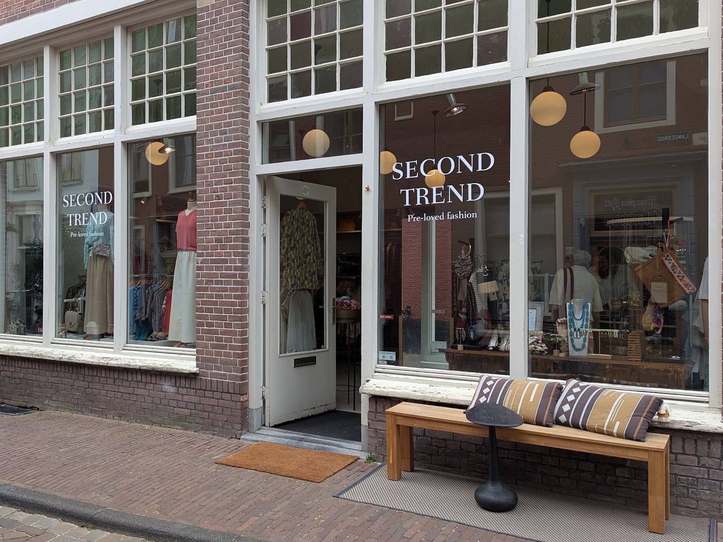 Bakstenen winkelpui van Second Trend met de naam op de ramen, een open deur en een bankje ervoor