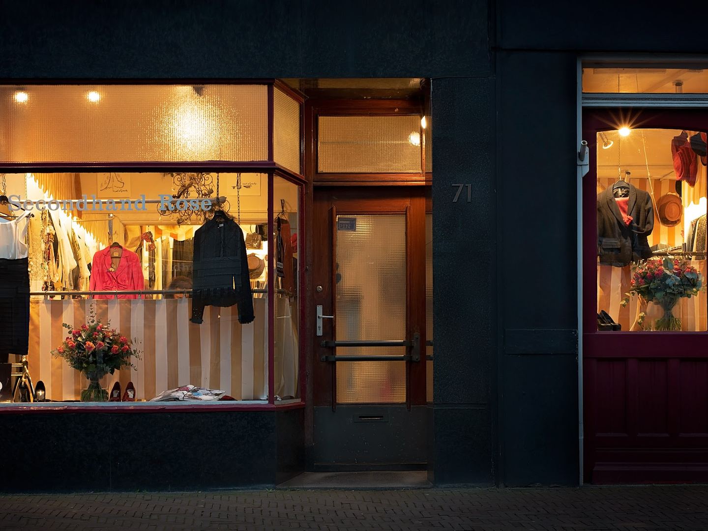 Donkere winkelpui van Secondhand Rose met twee verlichte etalages vol kleding en een houten deur ertussen