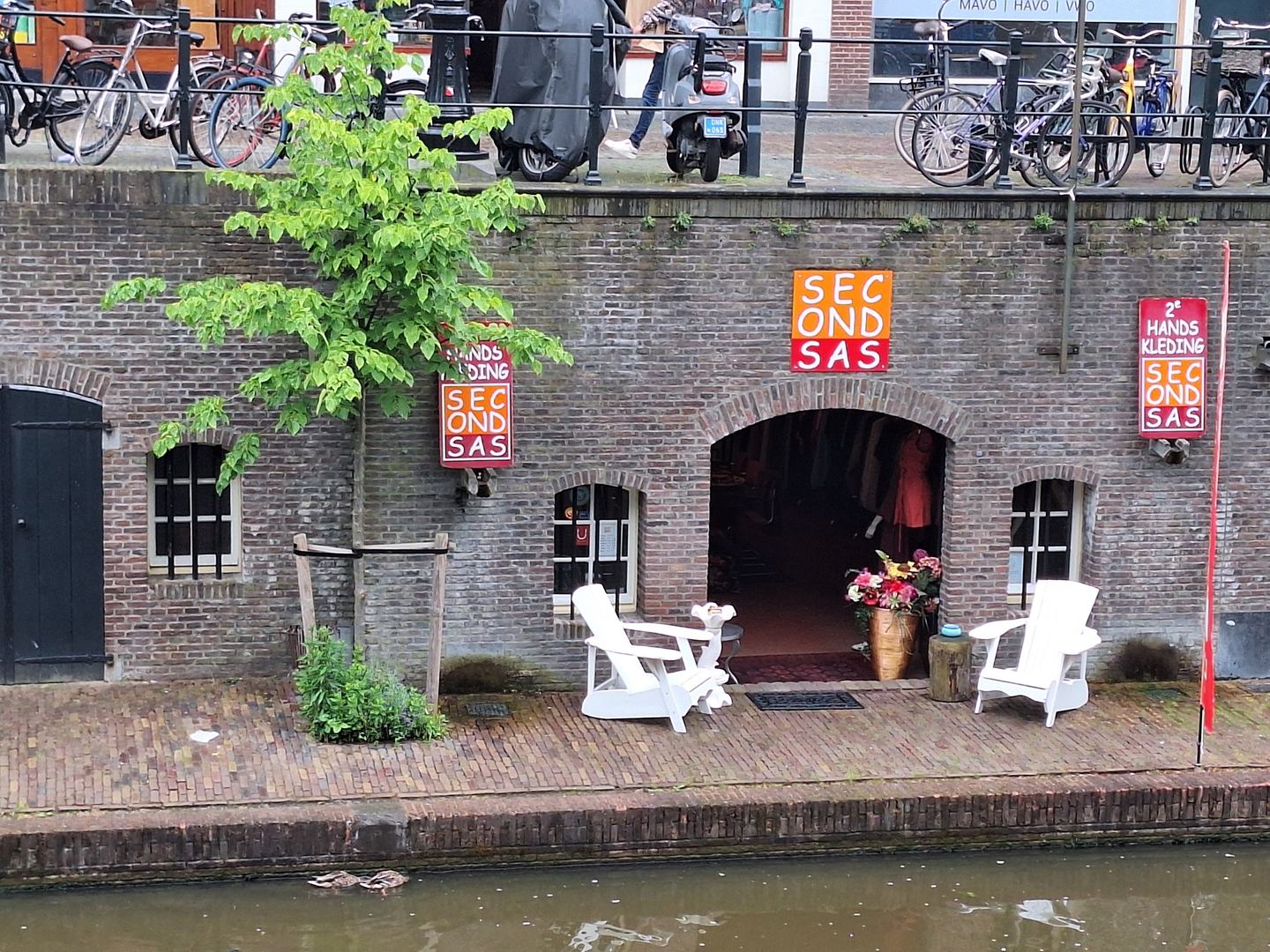 Ingang van SECONDSAS in een gemetselde werfkelder aan de gracht, met naamborden en witte tuinstoelen op de werf