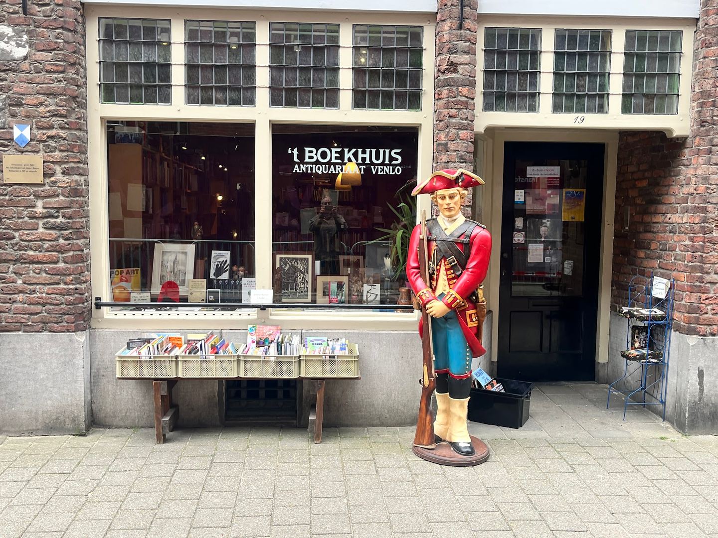 A storefront of 't Boekhuis' showing the window with the shop name, a book display, and a statue outside.