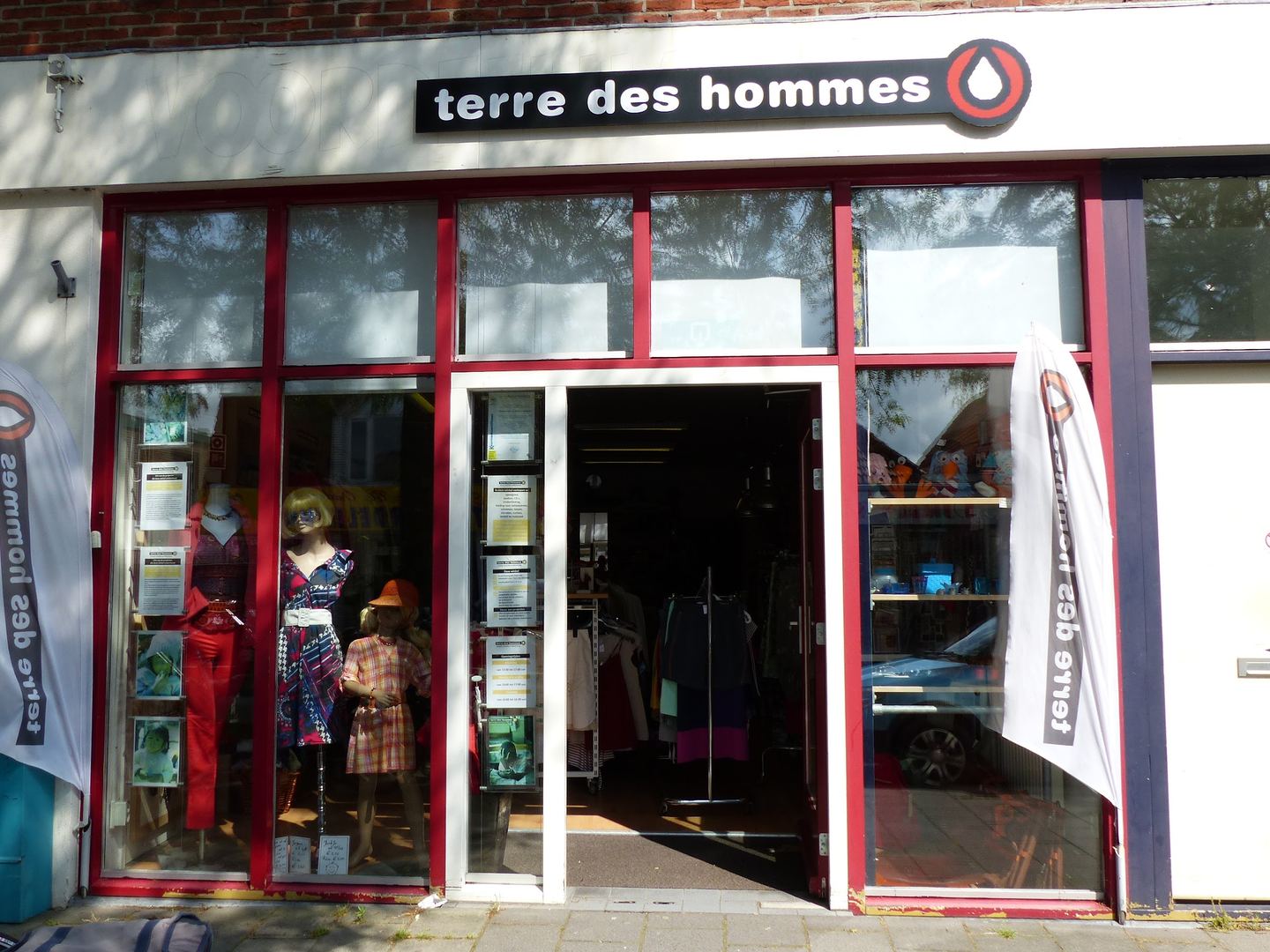 Winkelpui van Terre des Hommes met naambord, open deur, paspoppen in de etalage en twee banieren