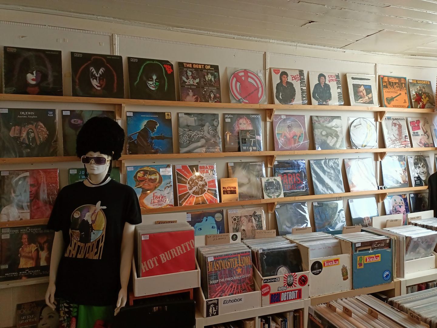 Wand vol platenhoezen in The Shop Records met bakken lp's eronder en een paspop in een bandshirt
