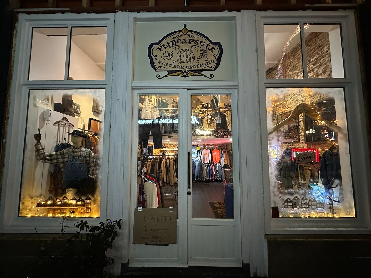 Witte gevel van Tijdcapsule met naambord boven de glazen deur en verlichte etalages vol vintage kleding