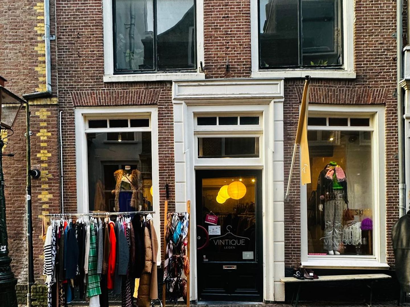Bakstenen gevel van Vintique met etalages, een open deur en rekken met kleding op de keien ervoor