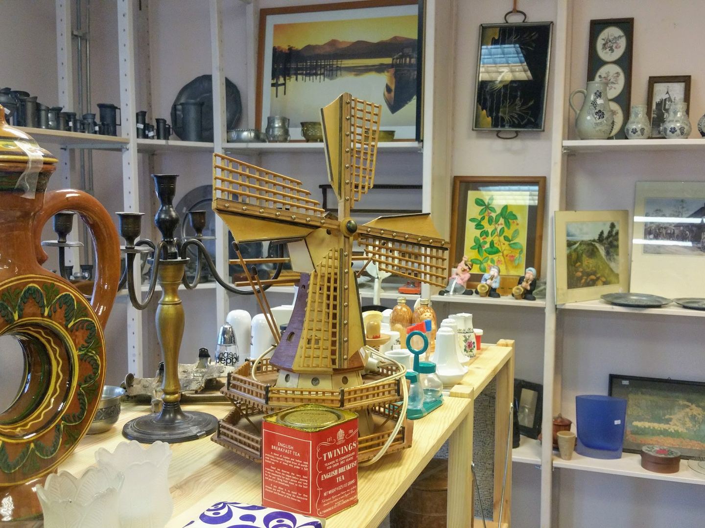Schappen met curiosa, tinwerk, kandelaars, schilderijtjes en een houten molen in de kringloopwinkel