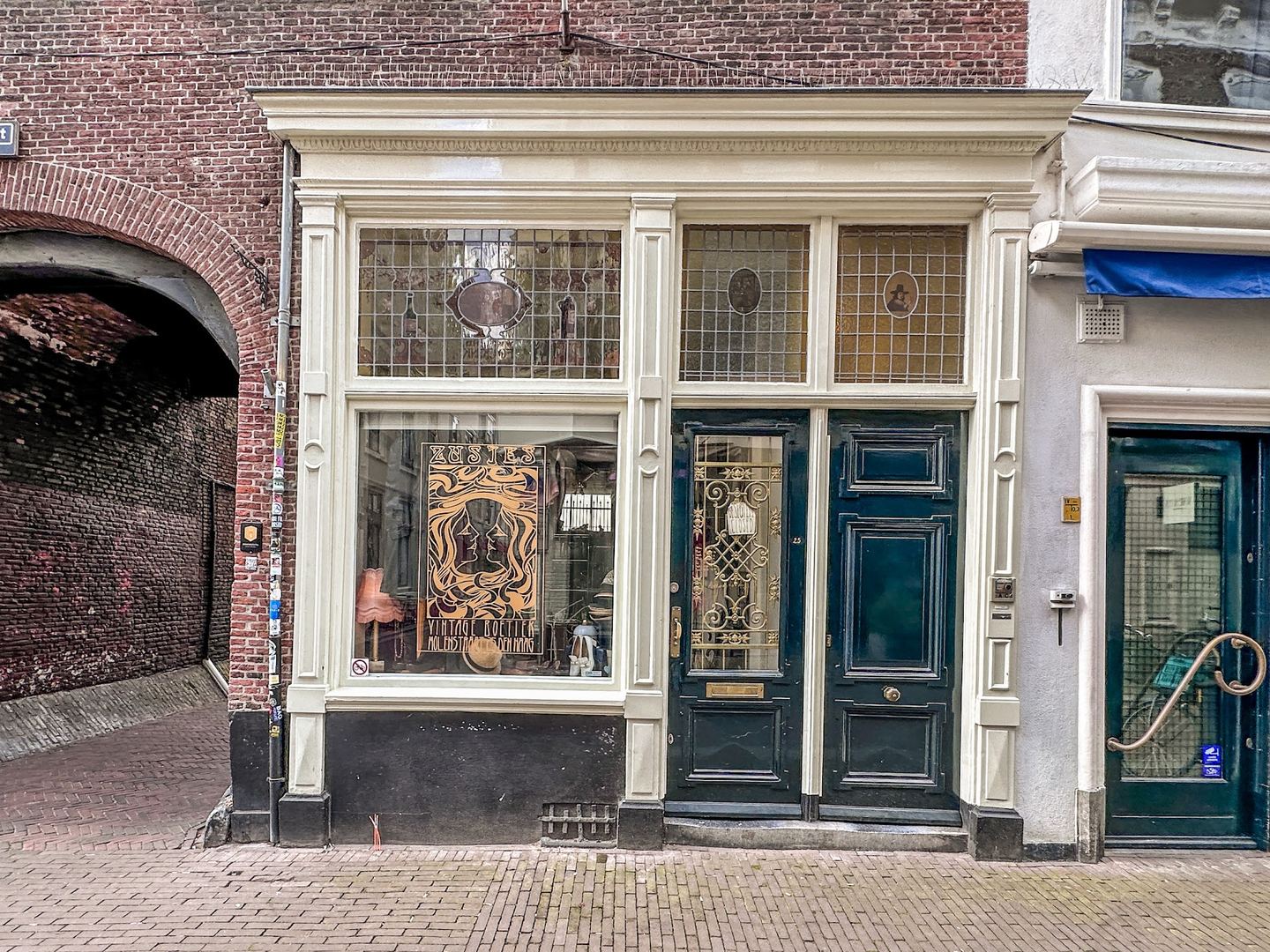 Gevel van ZUSJES vintage boetiek met glas-in-loodramen, een etalage en groene deuren naast een poortdoorgang